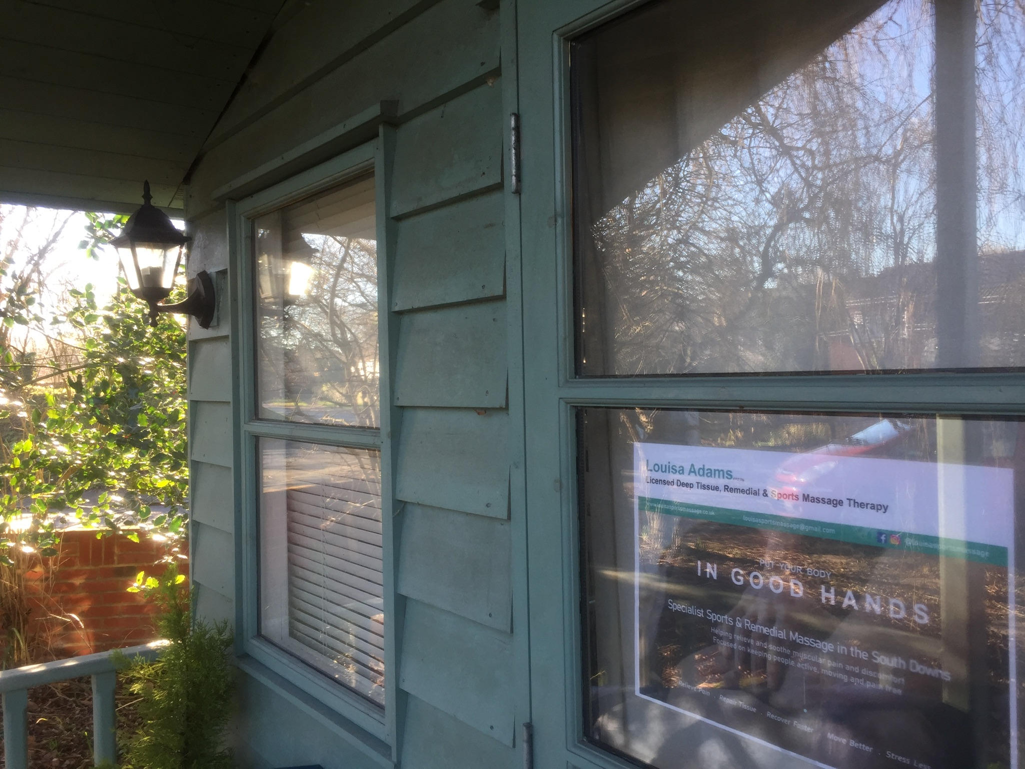 Louisa's studio: a sage-green painted timber cabin with a black lantern by the door and her poster in the window, winter sun coming through the trees.