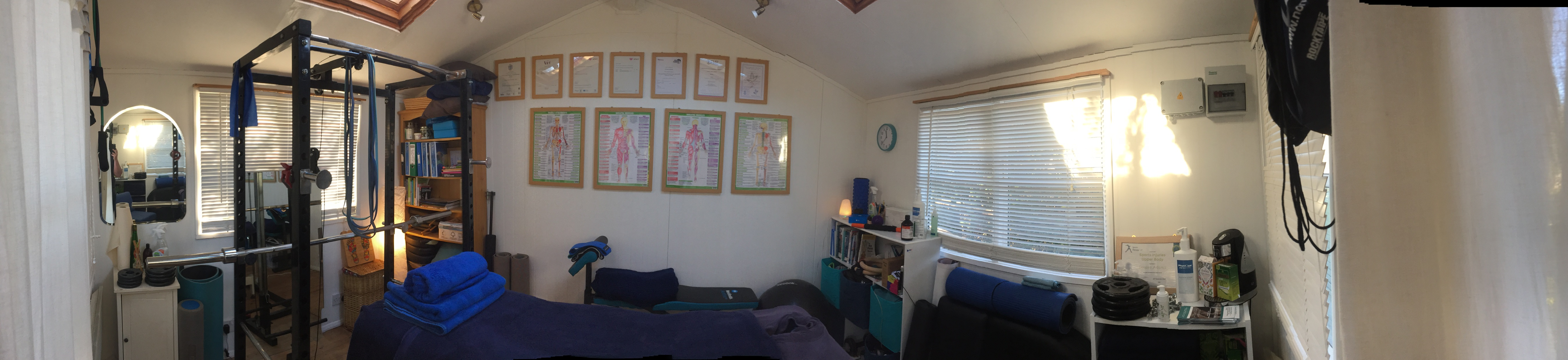 Wide panorama of the studio interior: treatment couch with blue towels, a squat rack, bookshelves, certificates and anatomy charts, and a window with the blinds half open.